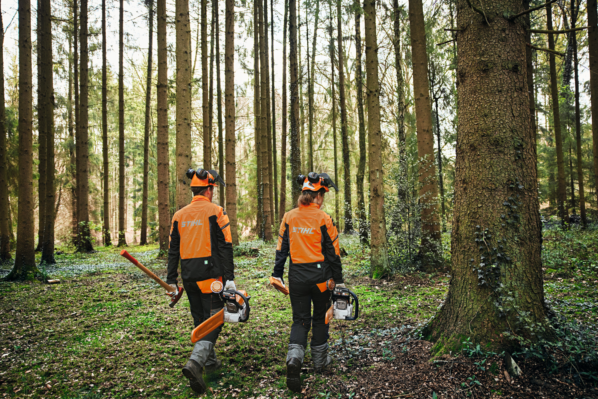 Két ember az erdőben védőfelszerelésben láncfűrészekkel és fejszével.