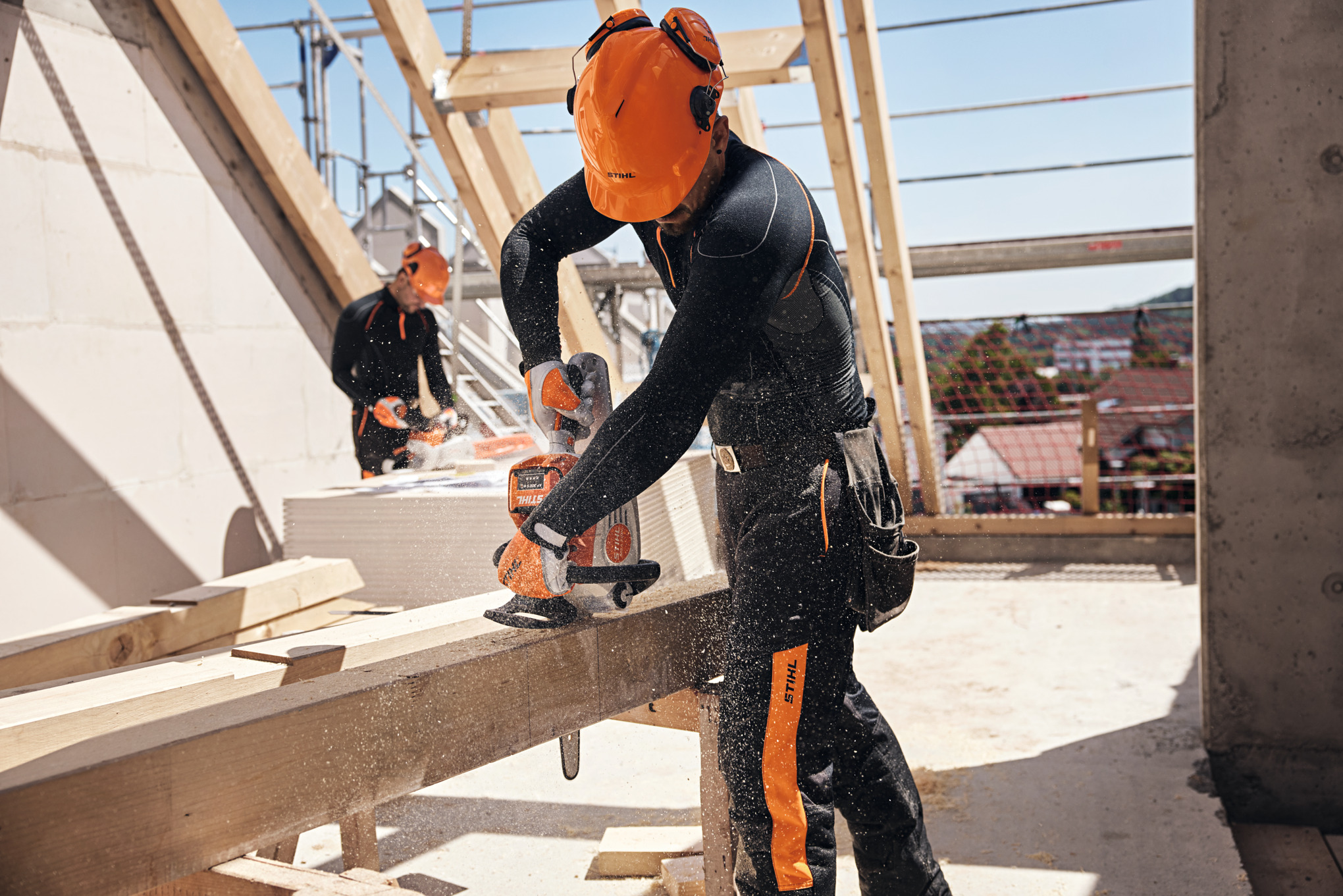 Egy STIHL szakember a tetőszerkezeten fűrészel egy akkumulátoros fűrésszel, a háttérben egy másik munkás akkumulátoros fűrésszel dolgozik.