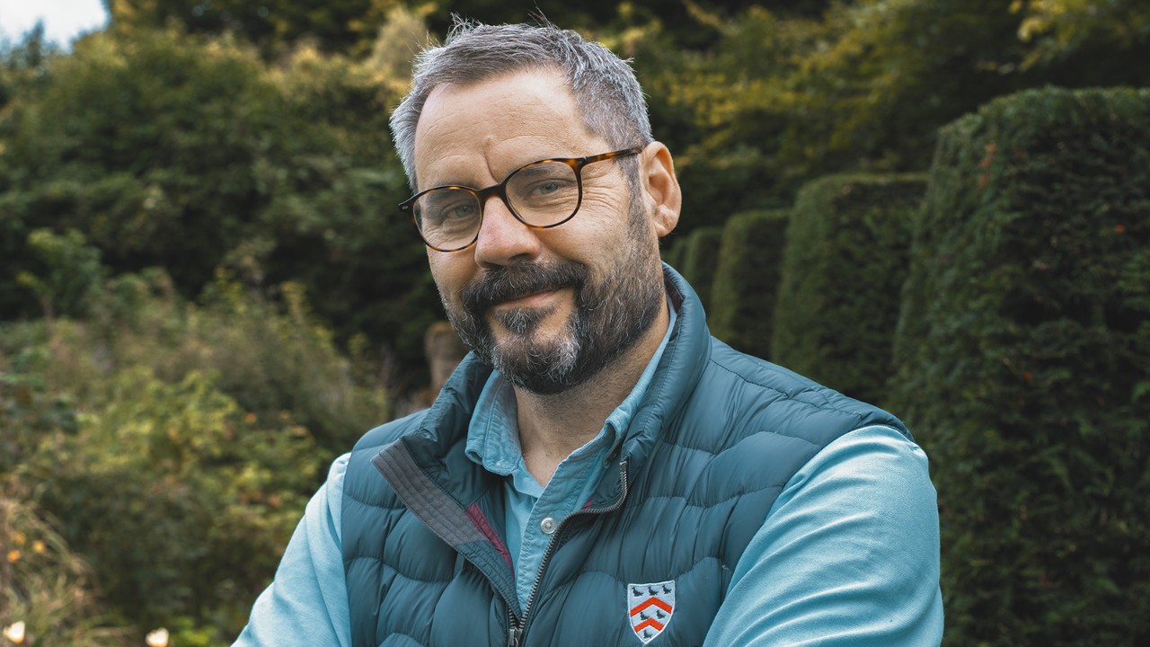 Portré a Worcester College kertészeti és kertészeti vezetőjéről, aki a szabadban áll egy parkosított kertben, szakmai munkaruhát és szemüveget visel.