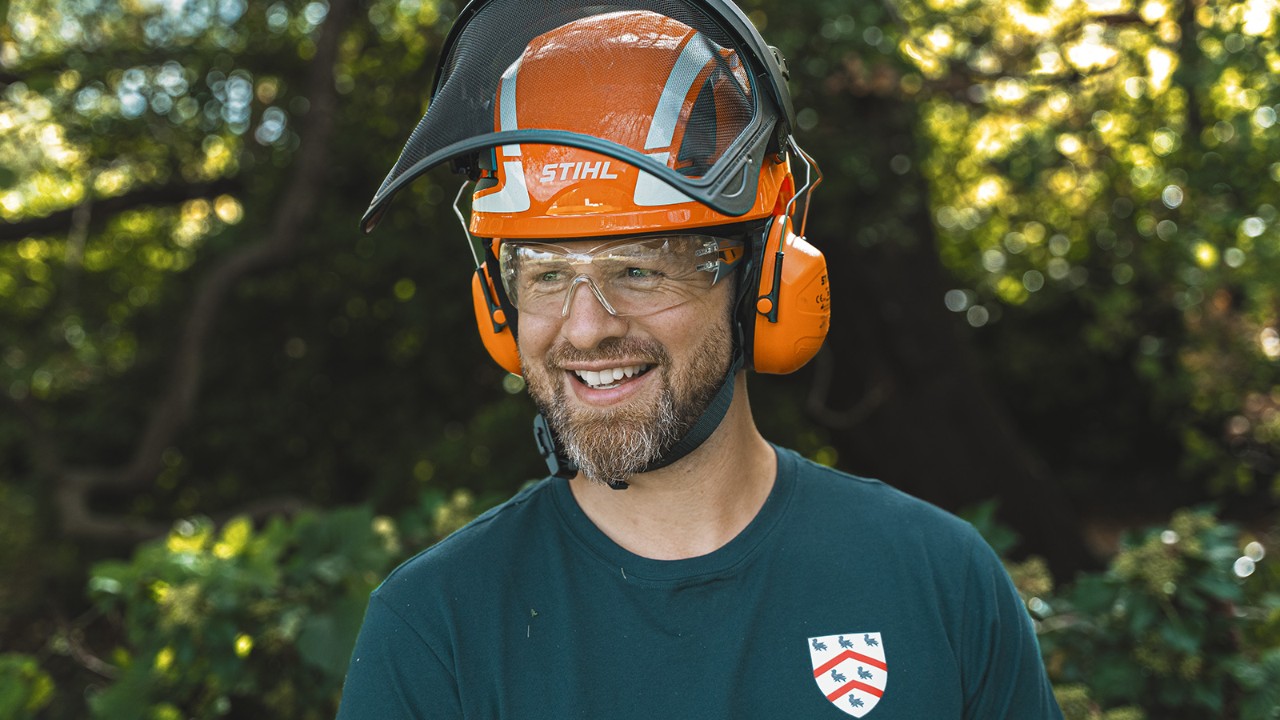 A Worcester College egyik arboristájának portréja, aki védő fejfedőt és munkaruhát visel, és a szabadban áll egy erdős környezetben.